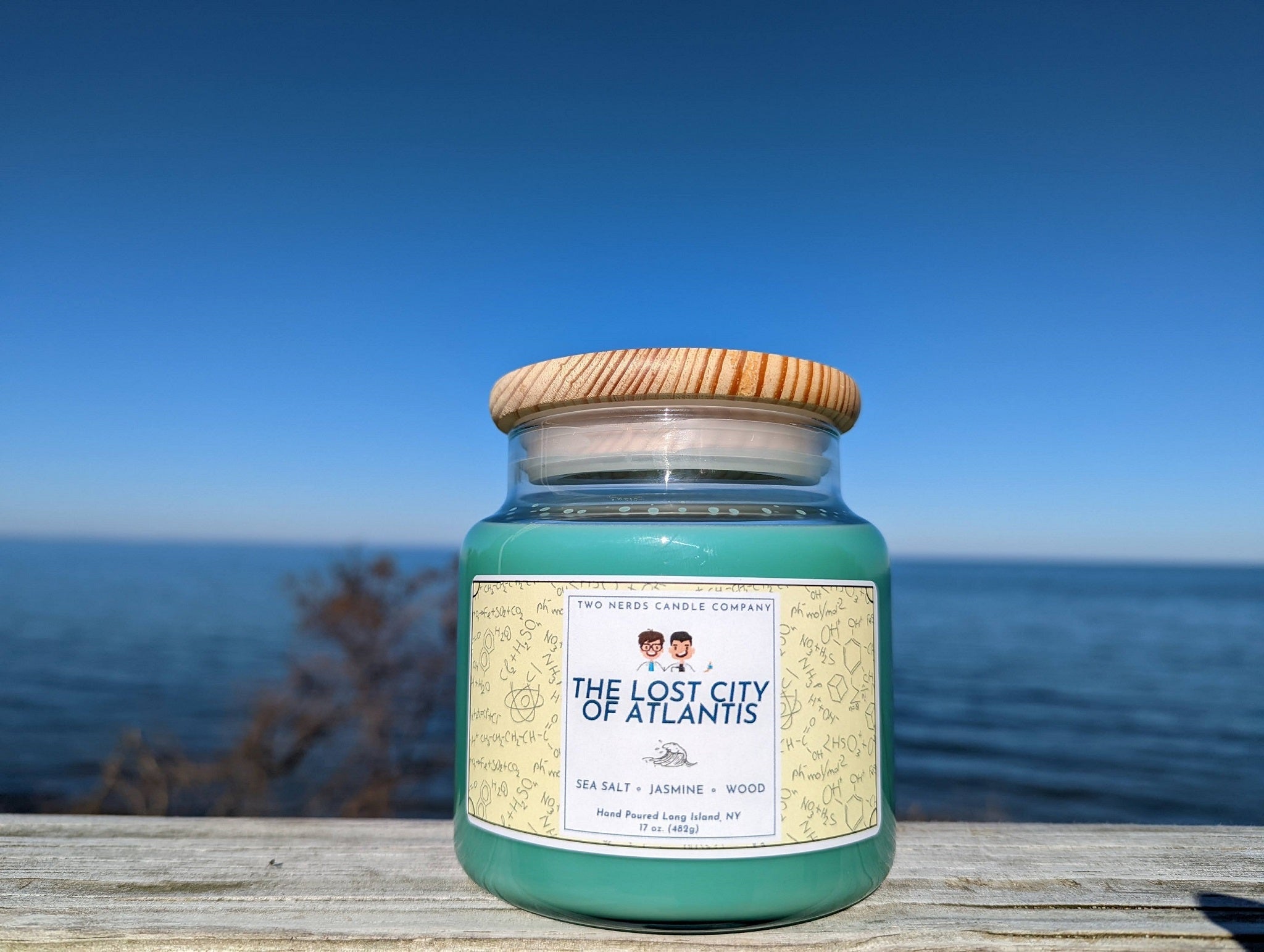 A beach themed soy candle which is placed on a beach background. The aqua colored candle is placed on a wood table with the ocean in the background.